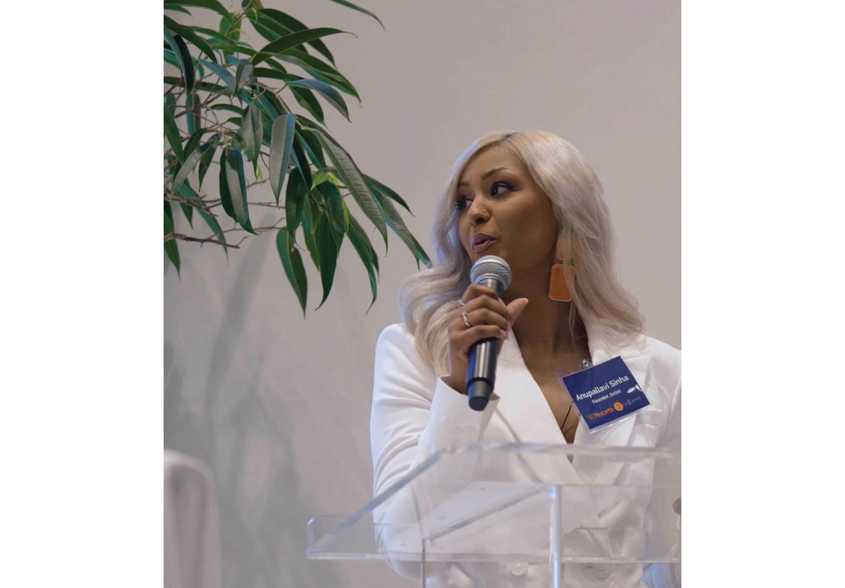 Anupallavi Sinha wears white and speaks into a microphone, alongside a green plant.