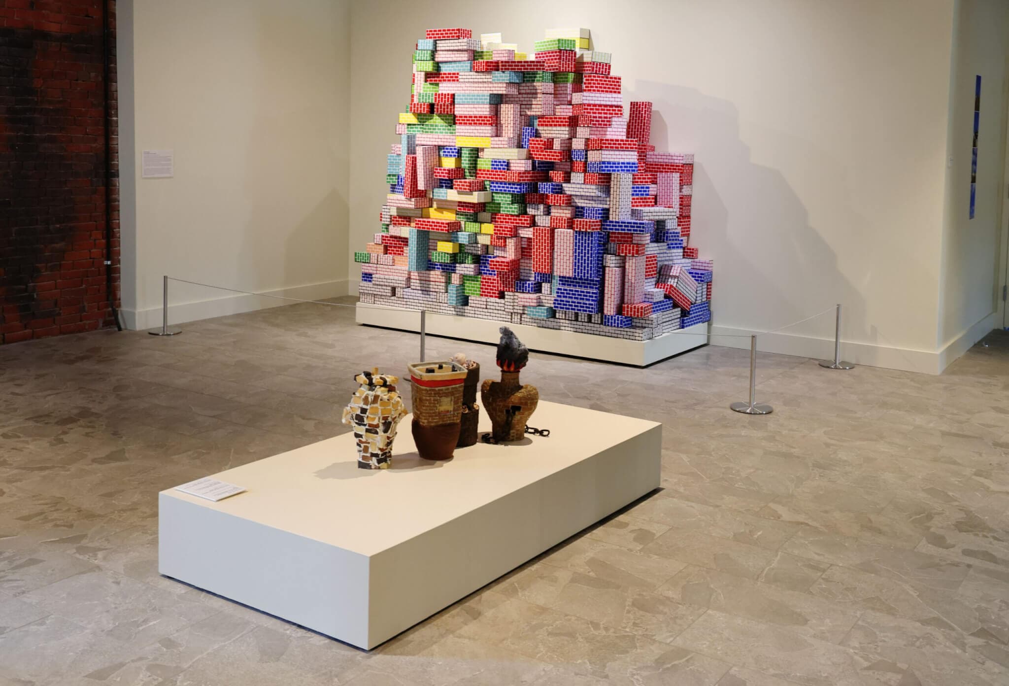 A sculpture made up of colorful cardboard bricks rests on a pedestal against a wall; small brick-based sculptures are presented on a pedestal at the center of the white-walled room.