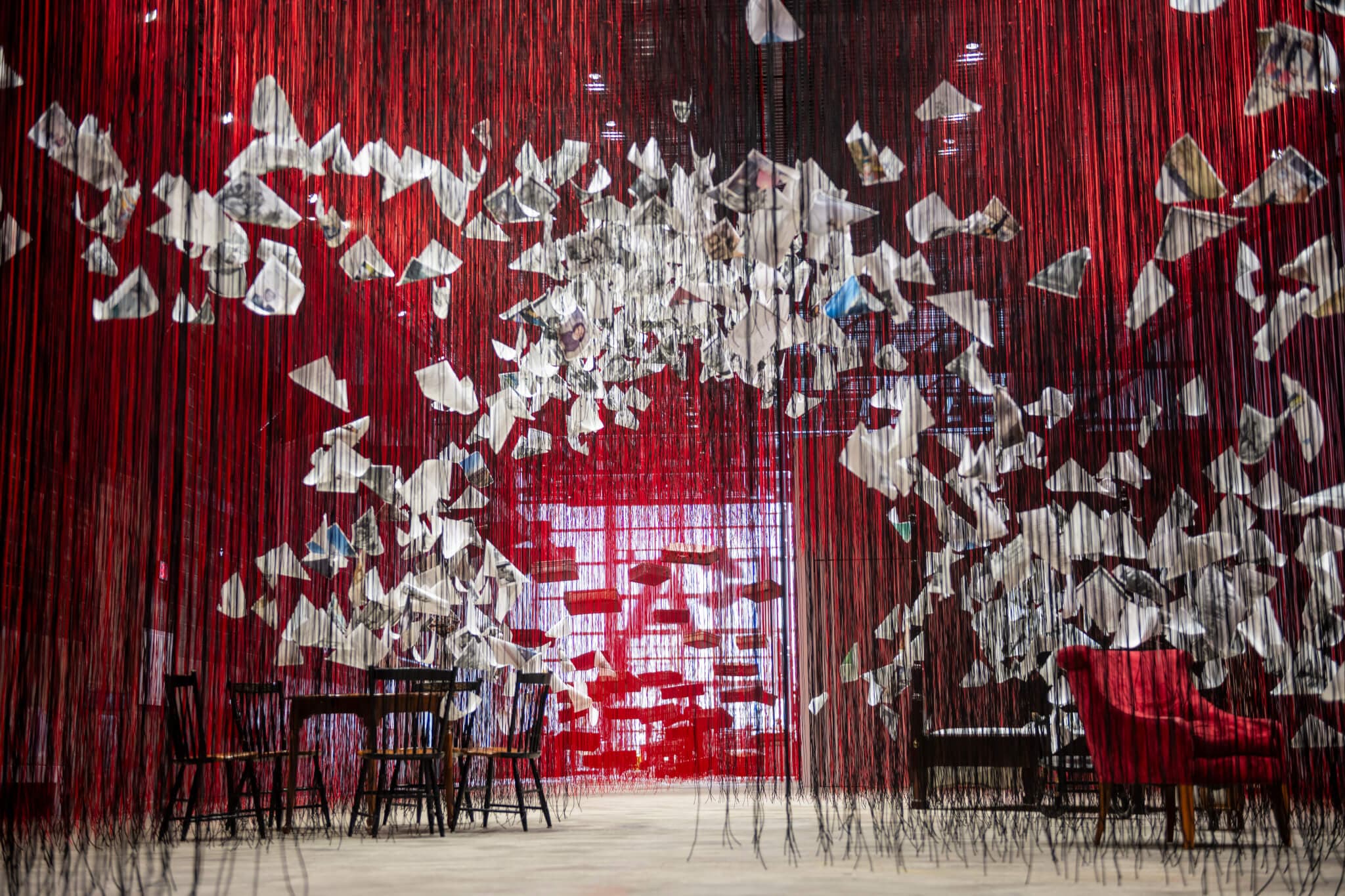 Papers dangle from red threads hanging from the ceiling of a large gallery space.