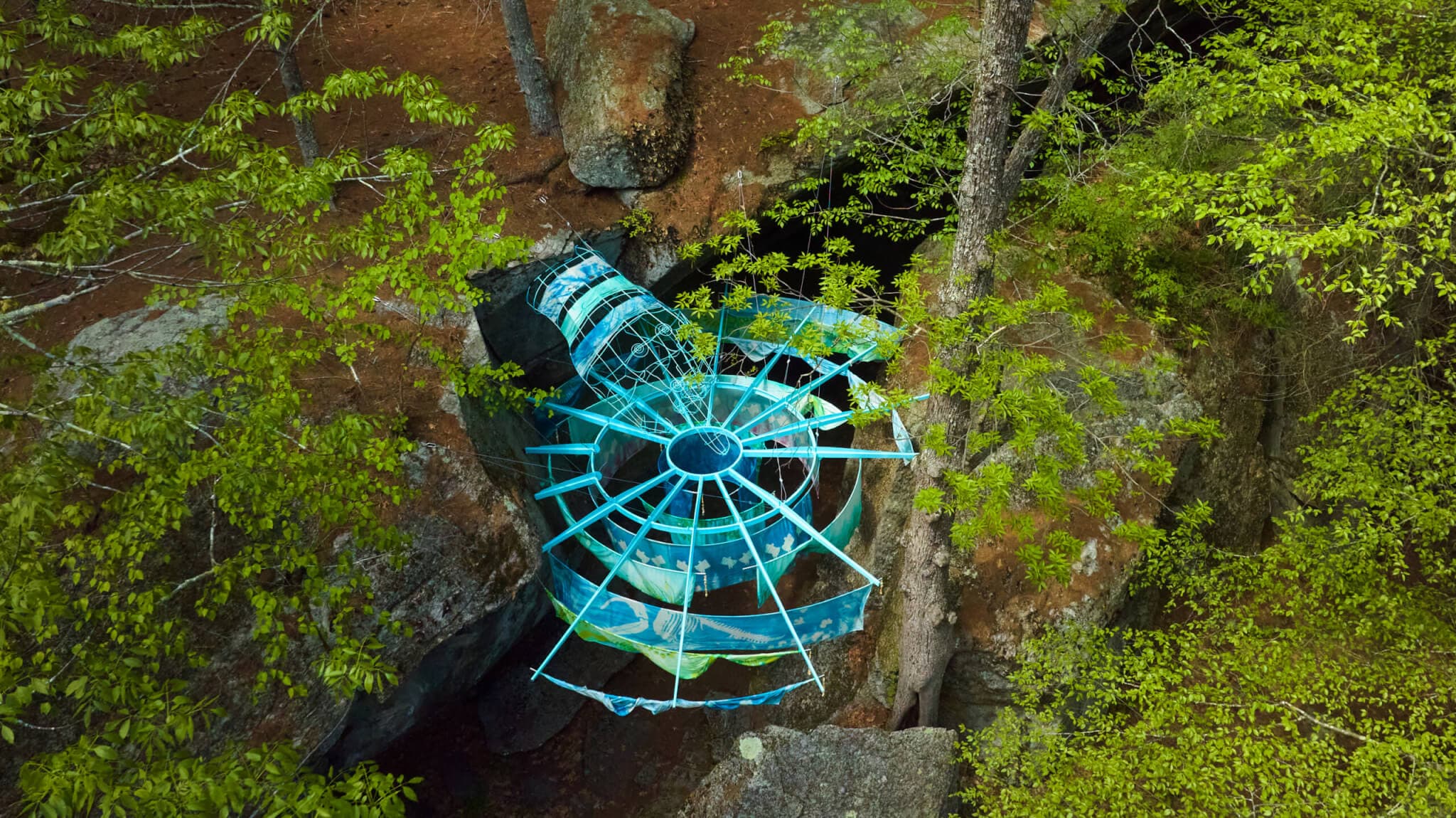 A blue metal sculpture with hanging silks is situated between rocks in a forest.